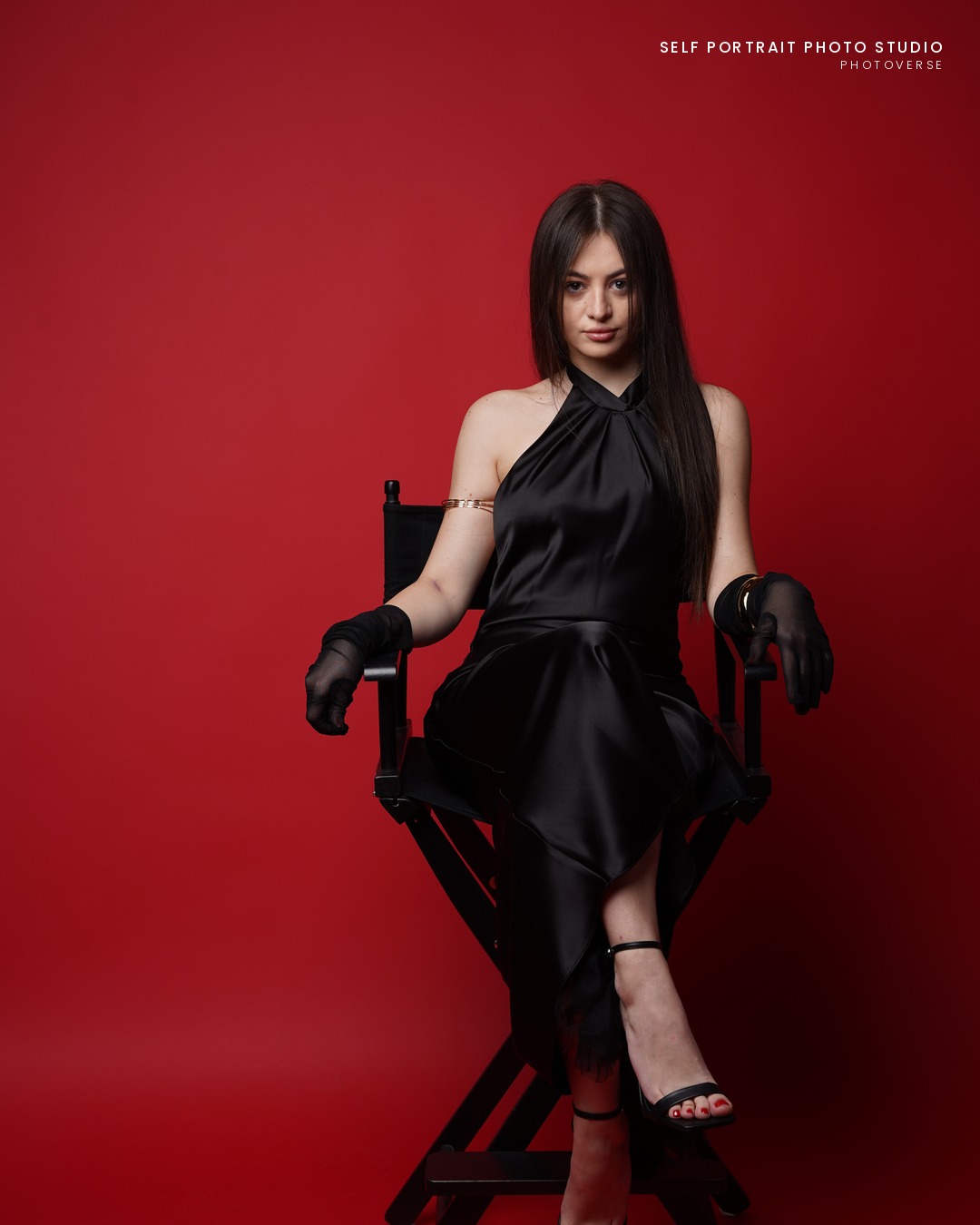 Self-portrait in a black gown seated on a director's chair, red backdrop
