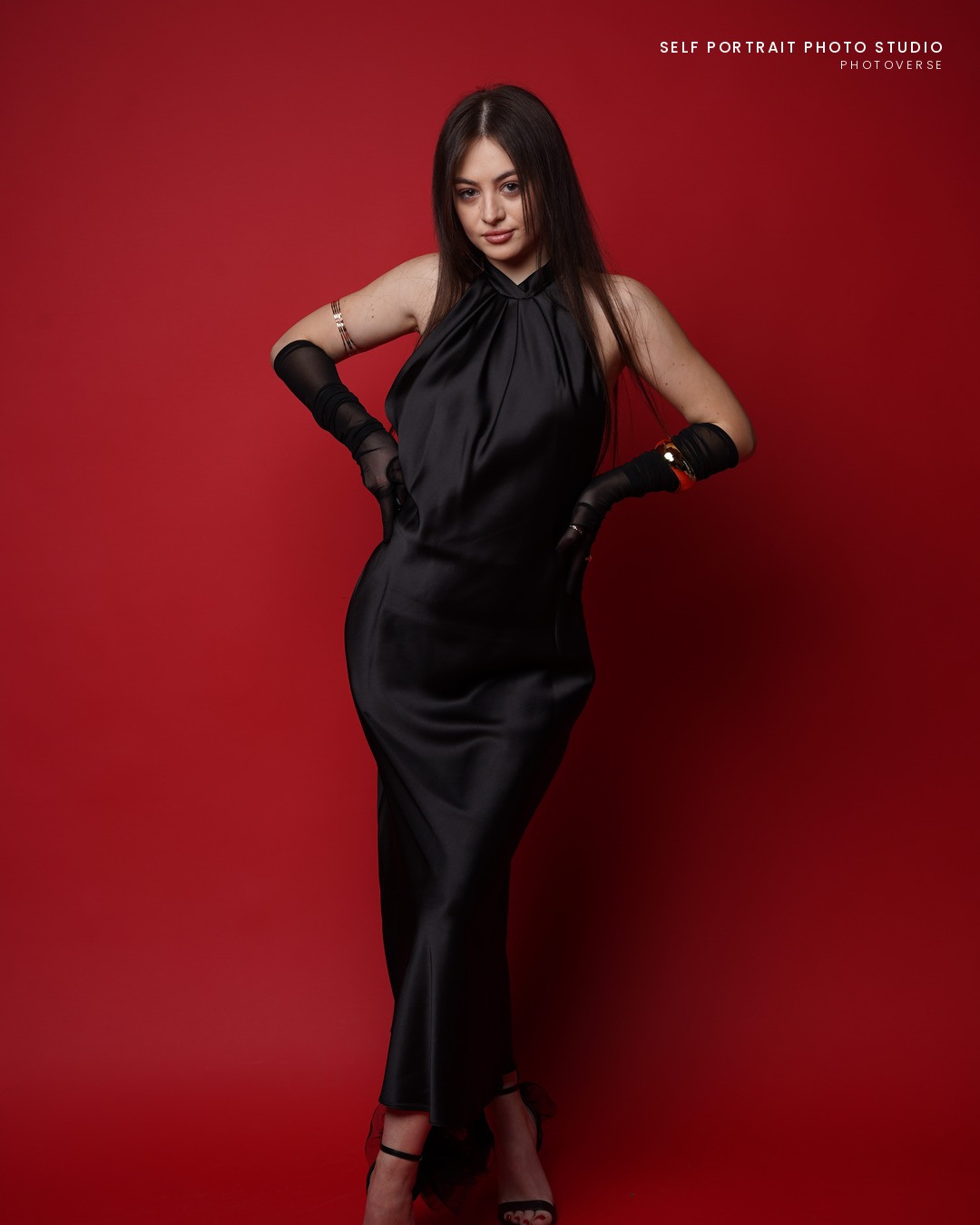 Close portrait in a black satin dress, hands on hips, red backdrop
